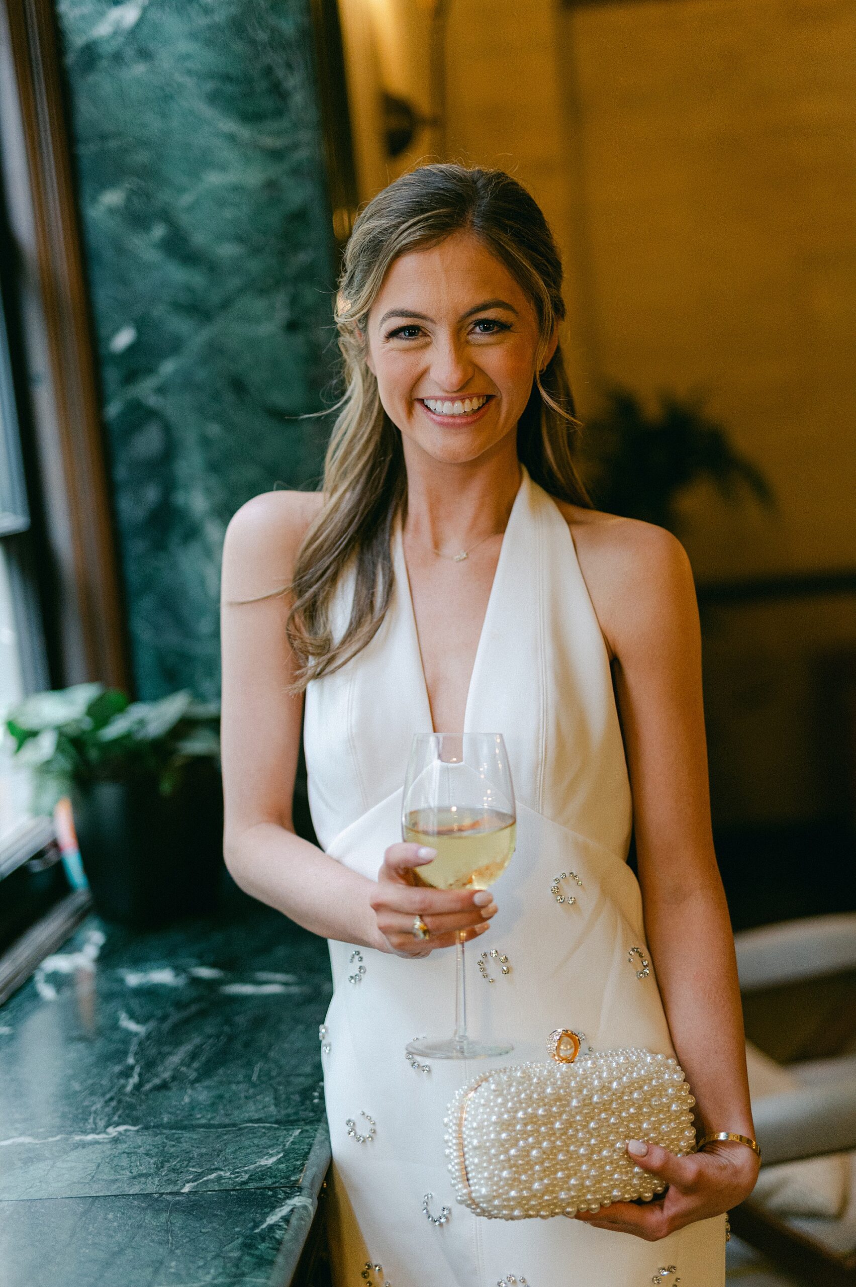 beautiful bride-to-be in white gown holding glass of wine and pearl clutch